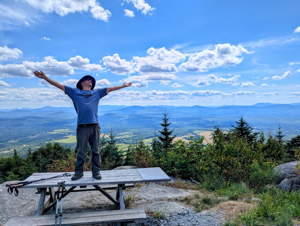 Carl Boileau au sommet d’une montagne les bras ouverts devant un vaste paysage du Québec.
